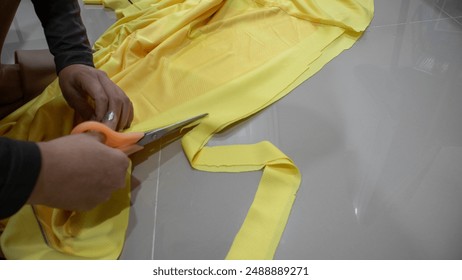 a persons hands using a pair of orange scissors to cut through yellow fabric. The fabric is spread out on a flat, reflective surface, possibly a table or counter. The hands are holding the fabric taut