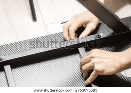 Person's hands work on assembling a black metal frame with a carbon fiber surface on a light wooden floor, with tools and parts nearby