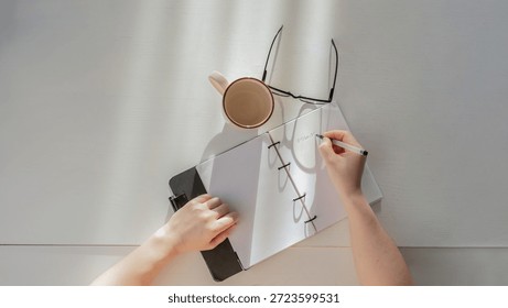 Person's hands writing notes in a planner next to a cup and glasses on a minimalist desk, top view