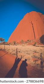 A persons shadow on the ground in front of a large red rock.
