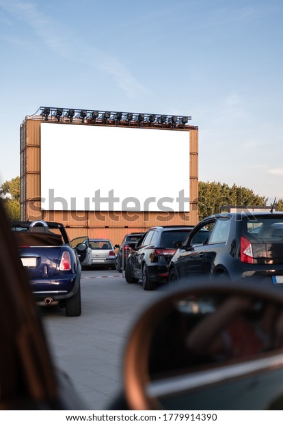 Perspective of a cinema drive in parking with\
cars parked in front of a big white screen to watch movies or films\
inside the car. Open air public Cinema drive-in. Social Distance,\
covid-19