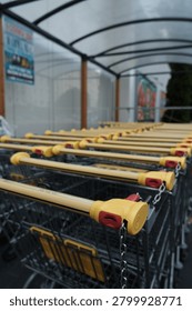 A perspective close-up of a long row of empty metal shopping carts nested together outside a supermarket. The shallow depth of field sharply focuses on the bright yellow plastic handles equipped with red coin-operated locks and connecting metal chains in the foreground. The background features a softly blurred outdoor cart shelter with a curved translucent roof and store advertisements, highlighting an everyday retail and grocery shopping environment