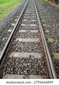 A perspective close-up view of a train railway track showing steel rails, concrete sleepers, and dark gravel ballast stones after rain