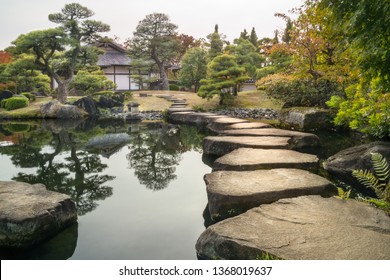 Perspective With Large Flat Rocks Forming A Path Over The Pond And With A Stone Lantern And A Japanese Building In Background, In Autumn, At Koko-en Garden In Himeji Located Next To The Famous Castle.