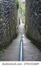 A perspective looking down a steep, narrow outdoor staircase with a central handrail, flanked by rugged, dark stone walls, leading to a path below in Dyserth, North Wales.