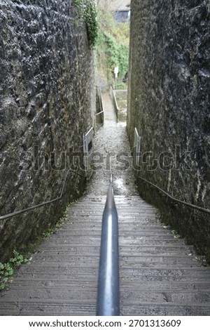 A perspective looking down a steep, narrow outdoor staircase with a central handrail, flanked by rugged, dark stone walls, leading to a path below in Dyserth, North Wales.