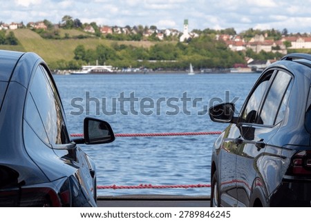 A perspective shot from the car deck of a ferry crossing Lake Constance (Bodensee). The camera is positioned between two parked cars, looking out towards the open water. In the distance, the hazy and soft-focus shoreline of Meersburg is visible under a bright, overcast sky