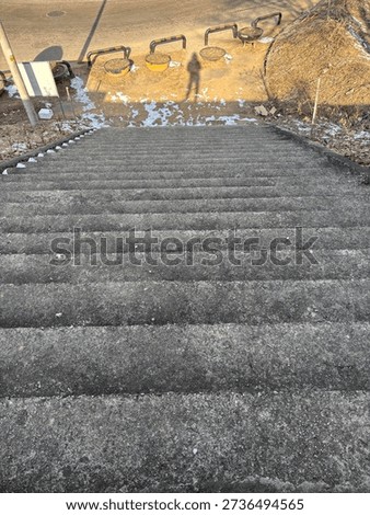 A perspective shot looking down a flight of gray concrete stairs, featuring the long shadow of a person cast by the bright sun onto the ground below.