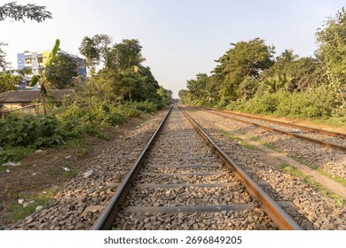 A perspective shot of parallel train tracks stretching into the distance, bordered by dense green foliage and trees. The sky above is clear and bright.