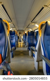 The perspective shot shows rows of empty blue seats with yellow grab handles inside a passenger train carriage, illuminated by overhead lights