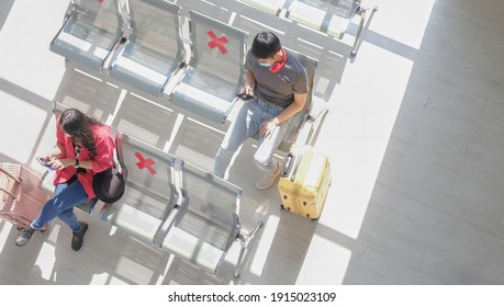 Perspective top view two young asian travelers wearing protective face mask, using smartphone while sitting follow social distancing regulation at public terminal station. COVID-19 awareness concept.