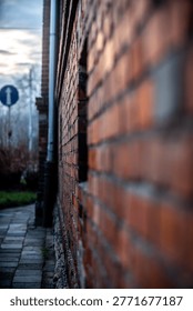 Perspective view of an aged red brick wall of an industrial building in the evening light with a blurred urban background, sidewalk, and a blue traffic sign under a cloudy sky.