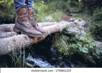 perspective view closeup of woman with brown tall leather hiking boots and grey jeans standing on a wooden trunk tree bridge over a mountain water stream in the forest - Powered by Shutterstock