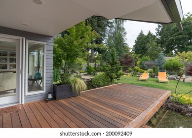 A perspective view of a contemporary Pacific Northwest home with a deck bridging a pond that leads to a pair of modern yellow loungers in a landscaped yard. 