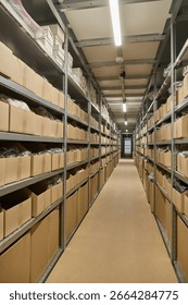 Perspective view down a long, narrow aisle in a large warehouse, flanked by towering metal shelving units densely packed with cardboard boxes. Industrial storage.