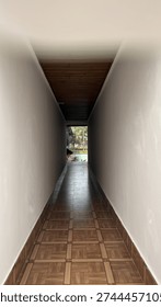 A perspective view down a long, white walled corridor with a tiled floor, opening up to a bright outdoor area with lush plants.