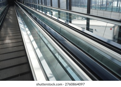 A perspective view of an empty moving walkway or travelator in a modern airport terminal, featuring glass side panels and metallic architectural details.