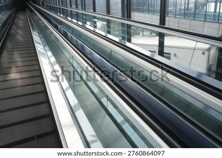 A perspective view of an empty moving walkway or travelator in a modern airport terminal, featuring glass side panels and metallic architectural details.