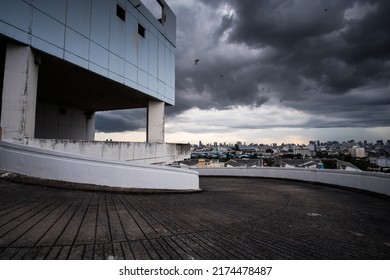 Perspective View Of An Empty Rooftop Car Park Of An Abandoned Mall, Dark Rain Clouds Cover A City In The Backgrounds.