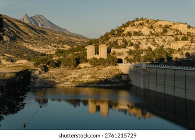 A perspective view of the massive concrete wall and green spillway gates of the Amadorio Dam in Alicante. The still reservoir water reflects the industrial scale of this civil engineering project. Arid mountains are visible above the structure. This facility is critical for water management and irrigation in the Valencian Community of Spain.