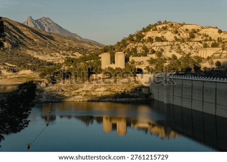 A perspective view of the massive concrete wall and green spillway gates of the Amadorio Dam in Alicante. The still reservoir water reflects the industrial scale of this civil engineering project. Arid mountains are visible above the structure. This facility is critical for water management and irrigation in the Valencian Community of Spain.