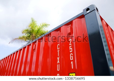 Perspective view of a red cargo shipping container with brown frame and white markings, background of pale cloudy sky and partial view of a palm tree top.