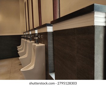 Perspective view of a row of white ceramic urinals with metallic flush valves mounted on dark tiled walls separated by elegant partitions in a modern male public restroom interior.