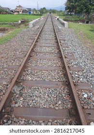 Perspective view of a rusty single-track railway passing through a tranquil rural landscape with lush green rice fields and distant mountains under an overcast sky. The train tracks stretch into the vanishing point, symbolizing travel, journey, transportation, and industrial infrastructure in the countryside