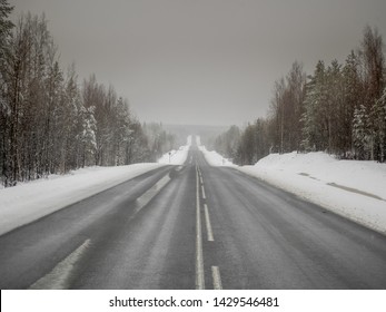 Perspective View Of The Straight Highway In The Winter Forest At Bad Weather. Dangerous Conditions For Driving. Icy Road With Snow Covered Roadsides And Coniferous Trees As Background. Symmetric View.