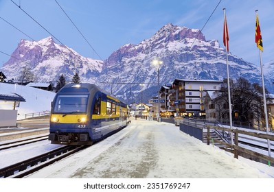 Perspective view of a train parking by a snow covered platform of Grindelwald Station on a cold winter evening, with majestic mountains in background at blue dusk, in Bernese Highlands, Switzerland