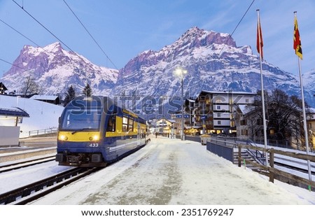 Perspective view of a train parking by a snow covered platform of Grindelwald Station on a cold winter evening, with majestic mountains in background at blue dusk, in Bernese Highlands, Switzerland