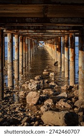Perspective view from underneath a rusty pier structure over shallow sea water with rocks. Strong leading lines and industrial texture concept. Perfect for travel, architecture, strength, and time passage themes.