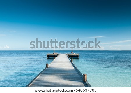 Perspective view of a wooden pier on the tropical seashore with clear blue sky with some white clouds and sea with turquoise water.