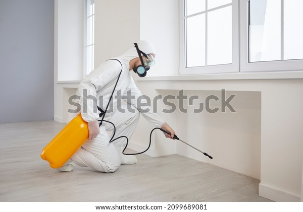 Pest control exterminator getting rid of insects\
inside infested house. Man in protective mask and suit spraying\
insecticide from yellow bottle on floor and walls for safe living\
environment at home