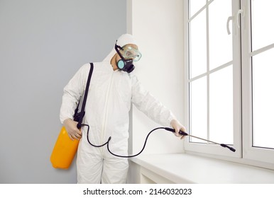 Pest Control Exterminator Working Inside House. Young Guy Wearing Protective White Suit And Mask Holding Sprayer Bottle And Spraying Insecticide Over Window Sill In Modern Living Room Interior At Home