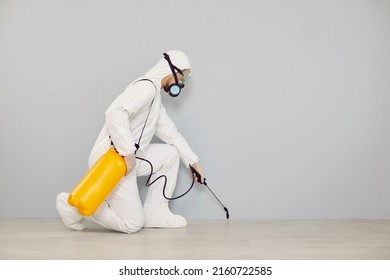 Pest Control Guy Cleaning Home From Insects. Man In White Protective Suit Crouching Near Wall And Spraying Floor With Cockroach Insecticide From Yellow Bottle For Safe Living Environment Inside House