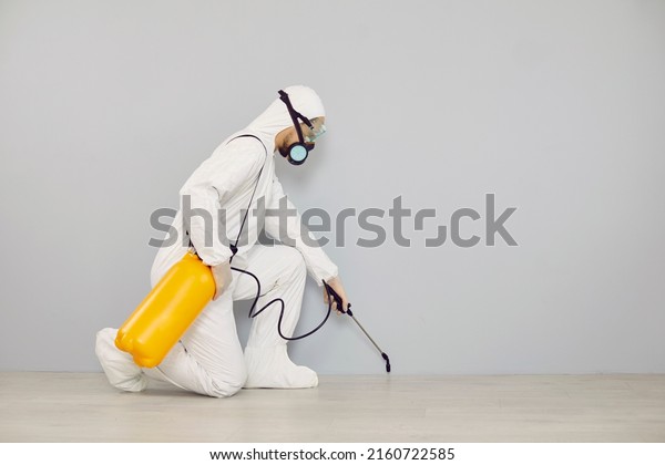 Pest control guy cleaning home from insects. Man in\
white protective suit crouching near wall and spraying floor with\
cockroach insecticide from yellow bottle for safe living\
environment inside house
