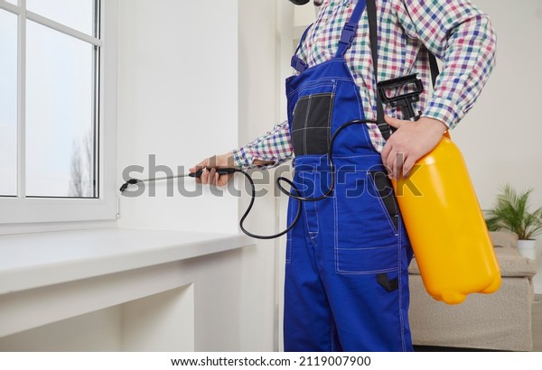 Pest control guy spraying poisonous gas or liquid\
on a windowsill at home. Male exterminator in overall uniform\
workwear spraying insecticide over a modern white window and sill\
inside the house
