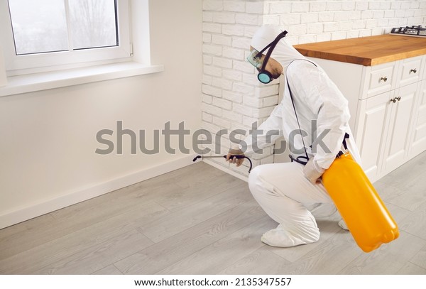 Pest control guy taking care of all the\
cockroaches or termites in the house. Male exterminator in white\
protective work uniform crouching by kitchen wall and spraying\
insecticide from sprayer\
bottle