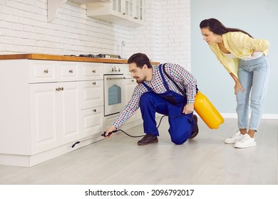 Pest control guy together with home owner spraying cockroach insecticide or rodenticide inside the house. Worker in blue uniform overalls crouching on floor and spraying chemical under kitchen cabinet