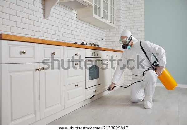 Pest control home service guy fighting parasites\
in the house. Exterminator in mask, goggles and PPE suit spraying\
poisonous gas or liquid from sprayer bottle on floor and cupboard\
in kitchen interior