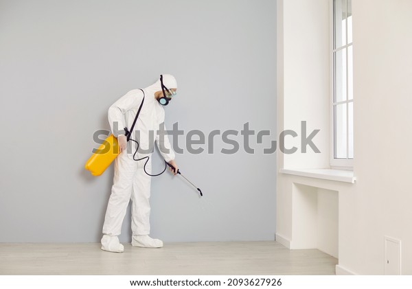 Pest control service guy visits the house. Man\
in white protective overalls holding yellow sprayer and spraying\
cockroach insecticide along wall for safe living environment inside\
residential building