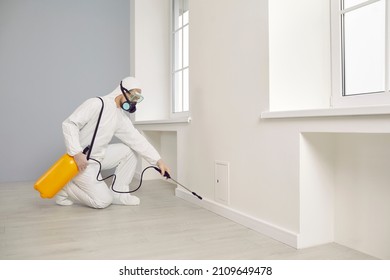 Pest control service guy working inside the house. Man in white protective overalls crouching near wall and spraying cockroach insecticide from yellow sprayer for safe living environment at home - Powered by Shutterstock