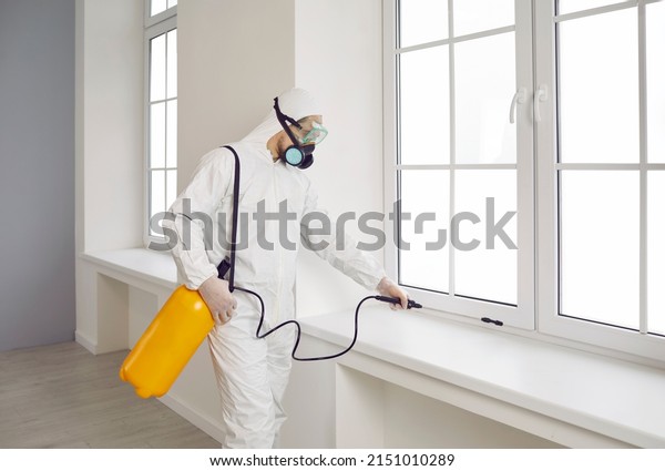 Pest control worker inside the house. Exterminator\
wearing white protective suit, mask and goggles holding yellow\
sprayer bottle and spraying insecticide over window sill in modern\
living room at home