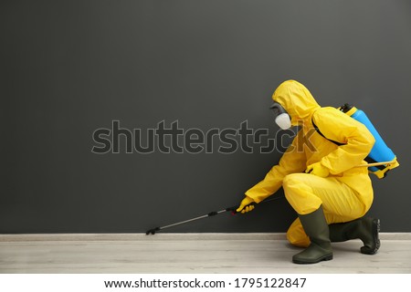 Pest control worker in protective suit spraying pesticide near black wall indoors. Space for text