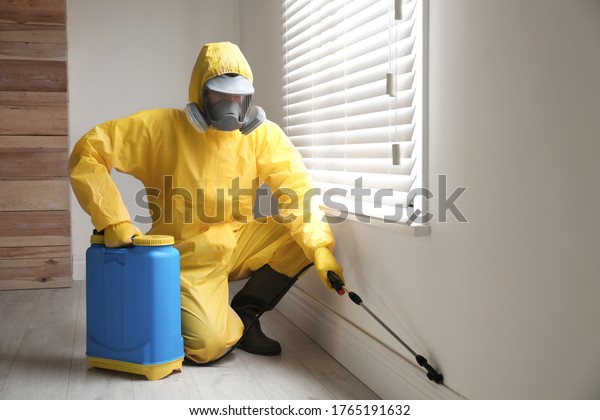 Pest control worker in protective suit spraying\
pesticide near window\
indoors