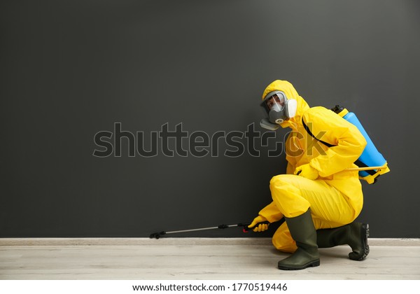 Pest control worker in\
protective suit spraying pesticide near black wall indoors. Space\
for text