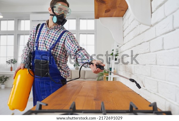 Pest control. Worker of pest control service\
during sanitary treatment of kitchen sprays poison. Man in goggles\
and respirator sprays on countertop insecticidal chemical spray\
from large spray bottle.