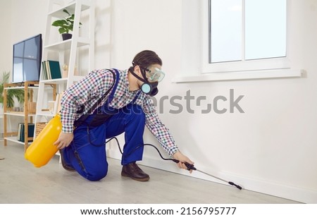 Pest control worker spraying insecticide in an infested home. Young male exterminator wearing a protective mask and overall uniform disinsecting a modern living room interior of a new house