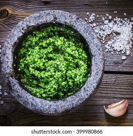 Pesto in marble mortar, salt and garlic on wooden table. Selective focus.